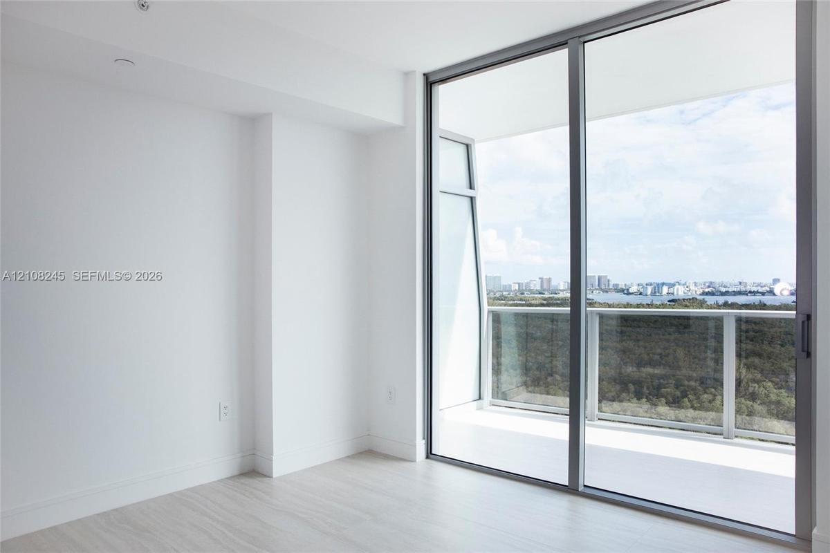 Floor-to-ceiling glass sliding doors, wood-finish flooring, and a private balcony featuring a glass railing and urban skyline scenery
