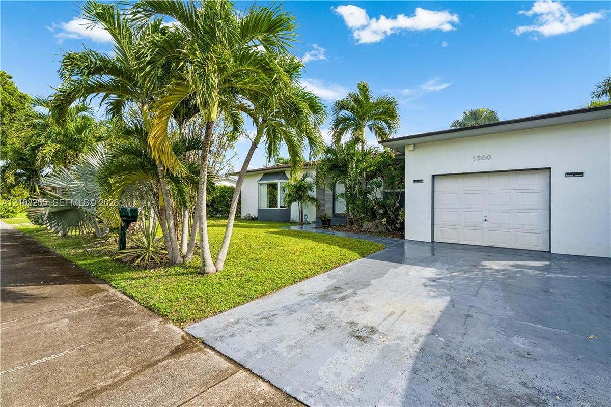 Single-story exterior featuring white stucco siding, a paved driveway, a one-car garage, and established palm landscaping