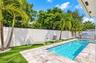 Private backyard featuring a rectangular swimming pool, stone paver decking, synthetic turf, mature palm trees, and a built-in outdoor kitchen