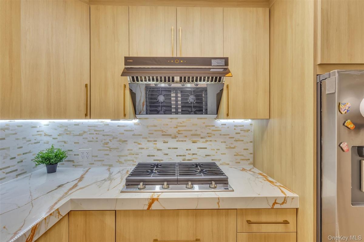 Kitchen featuring light wood-finish cabinetry, stone-patterned countertops, a tile backsplash, a stainless steel gas cooktop, and a matching range hoo