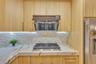 Kitchen featuring light wood-finish cabinetry, stone-patterned countertops, a tile backsplash, a stainless steel gas cooktop, and a matching range hoo