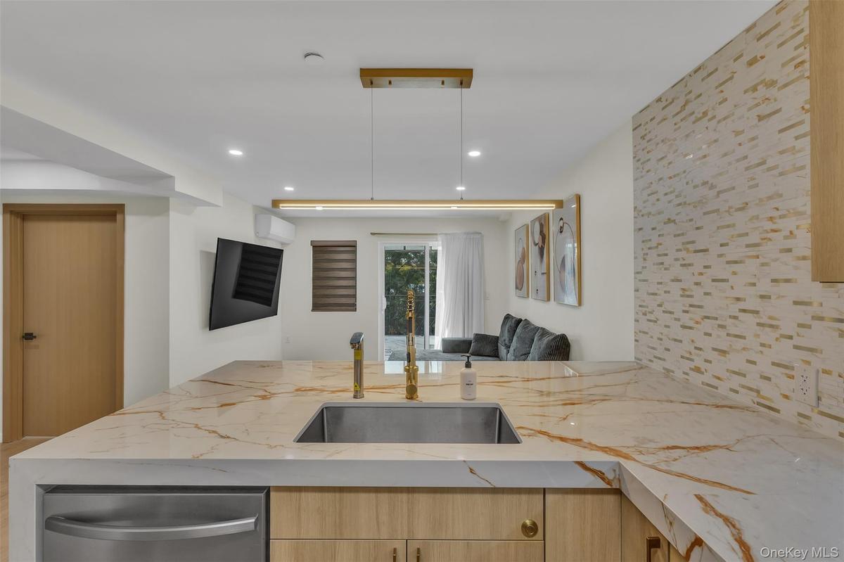 Kitchen island with stone-finish countertop, undermount stainless steel sink, gold-tone faucet, linear pendant lighting, and textured tile backsplash