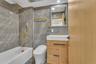 Bathroom featuring wall-to-ceiling large-format tile, vanity with wood-finish cabinetry, brass-tone plumbing fixtures, wall-mounted mirror, and a buil