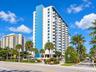 High-rise residential building exterior featuring a white and blue facade, prominent stacked balconies, and mature palm tree landscaping