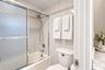 Bathtub with glass sliding doors, integrated handheld shower fixture, light-toned wall tile with decorative accent border, and stone-finish vanity cou
