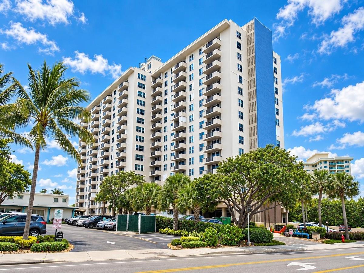 High-rise residential exterior featuring stacked private balconies, a distinctive vertical blue facade accent, and paved surface parking framed by mat