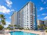 High-rise residential exterior featuring stacked private balconies, ground-level swimming pool, paved pool deck, and mature palm trees