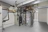 Mechanical room housing a forced-air furnace, industrial-grade water heater, metal ductwork, and exposed copper piping on concrete flooring