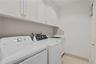 Laundry area featuring white shaker-style cabinetry, a stainless steel utility sink, tile backsplash, and light-toned tile flooring