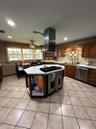 Kitchen featuring a central island with a built-in gas cooktop, stainless steel ventilation hood, wood cabinetry, stone-finish tile flooring, and rece