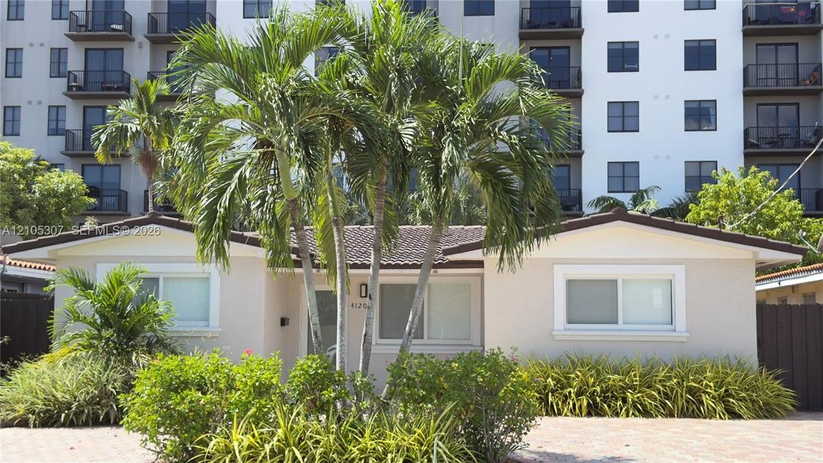 Single-story exterior featuring a light-colored stucco facade, tiled roof, paver driveway, and mature tropical landscaping