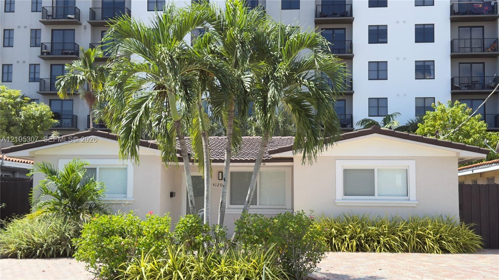 Single-story exterior featuring a light-colored stucco facade, tiled roof, paver driveway, and mature tropical landscaping