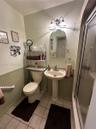 Pedestal sink with a dual-handle faucet, arched vanity mirror, wainscoting detail, light-toned ceramic tile flooring, and a glass-enclosed shower