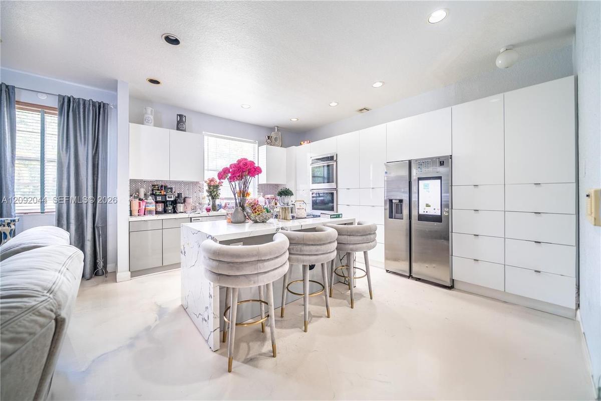 Modern kitchen featuring floor-to-ceiling high-gloss white cabinetry, a center island with stone-finish waterfall edges, recessed lighting, and stainl