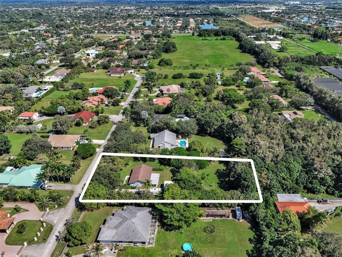Aerial view of a sprawling rectangular lot featuring extensive open green space, dense perimeter foliage, mature trees, and a single-story structure