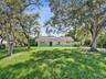 Single-story residence featuring a light-colored stucco exterior, a gable roof, and large mature shade trees situated on a spacious grass lawn