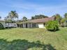 Single-story residence featuring a covered patio, shingle roof, exterior lighting fixtures, and a spacious grass yard with mature vegetation