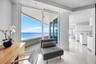 Floor-to-ceiling glass sliding doors opening to an expansive balcony with ocean scenery, white tile flooring, and sleek flat-panel cabinetry