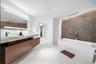 Floating wood-finish double vanity with stone countertop, large LED-backlit mirror, soaking tub, textured stone feature wall, and recessed ceiling lig
