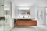 Floating wood-finish vanity with dual integrated sinks and stone-surfaced waterfall countertop, complemented by an illuminated vanity mirror and glass