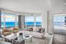 Floor-to-ceiling panoramic windows, expansive balcony with glass railings, polished tile flooring, recessed lighting, and coastal water backdrop