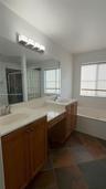 Dual-sink vanity with wood-finish cabinetry, solid-surface countertops, expansive wall mirror, and large-format tile flooring