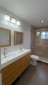 Dual-sink vanity with wood-finish cabinetry, rectangular under-mount basins, polished chrome fixtures, two wood-framed mirrors, and a glass-enclosed s