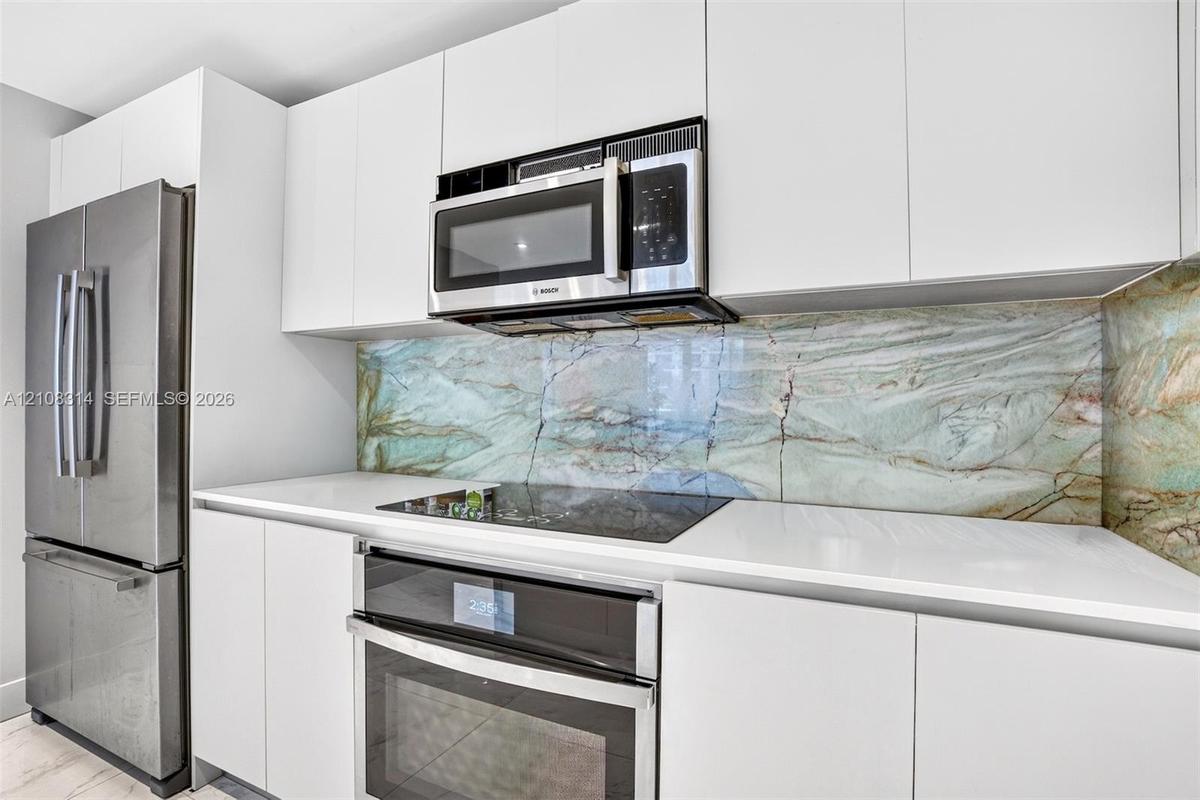 Kitchen featuring flat-panel white cabinetry, stone-patterned slab backsplash, quartz countertops, and stainless steel appliances including a built-in
