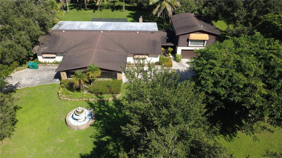 Aerial perspective of a multi-level residence featuring a multi-pitched roof, an attached garage, a stone-lined fountain, and expansive grounds with m