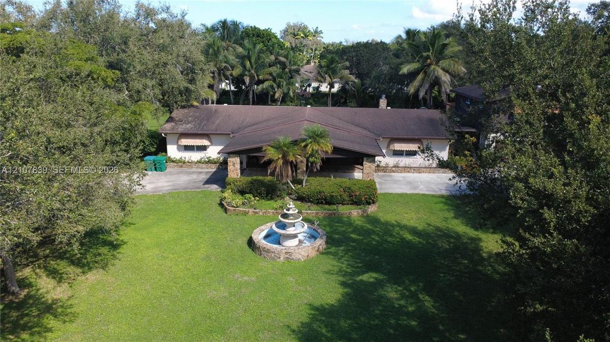 Single-story residence featuring a covered portico, stone-accented facade, central tiered water fountain, and expansive lawn surrounded by mature tree