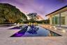 Infinity-edge pool featuring integrated lighting, expansive stone-paver patio, sliding glass door panels, and mature canopy trees