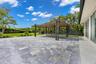 Spacious stone-paved patio featuring a covered pergola with recessed lighting and ceiling fans, manicured lawn, and privacy fencing