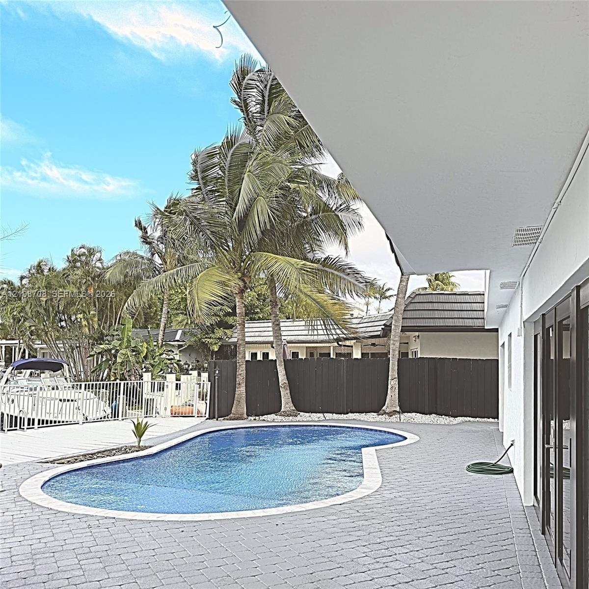 Backyard area featuring a kidney-shaped pool, grey paver decking, white metal fencing, and mature palm trees