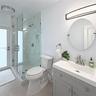 Glass-enclosed shower with stone-finish wall tile and pebble-texture flooring, grey shaker-style vanity with integrated sink, and circular wall-mounte