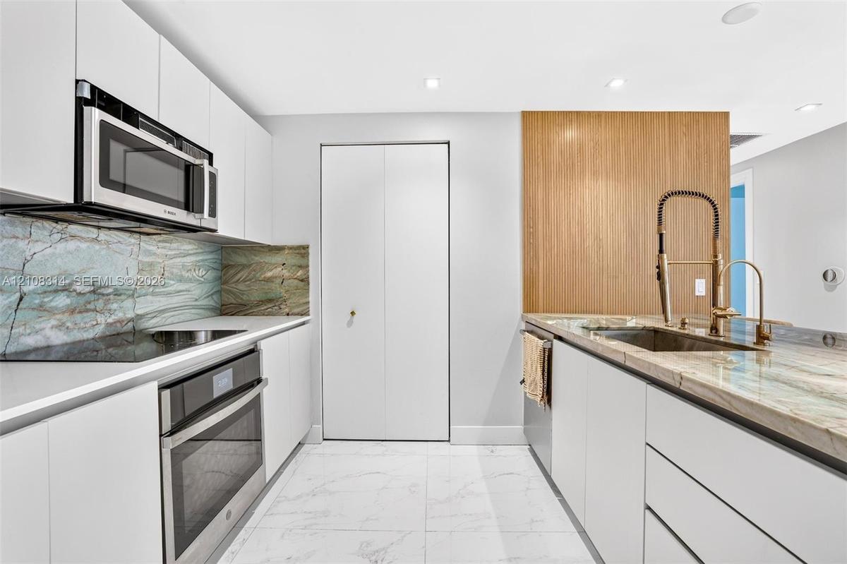 Modern kitchen featuring flat-panel white cabinetry, stone slab countertops, integrated stainless steel appliances, and wood-finish accent paneling wi