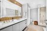 Dual-sink vanity with white surface, metallic gold-finish faucets, large-scale vanity mirror with integrated lighting, wood-slat wall paneling, and wa