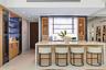 Custom bar area featuring stone slab backsplash, integrated wine refrigeration, built-in light wood cabinetry, stone-topped island with waterfall edge