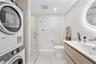 Stacked laundry units, walk-in shower with frameless glass enclosure, wall-mounted floating vanity with integrated sink, and illuminated vanity mirror