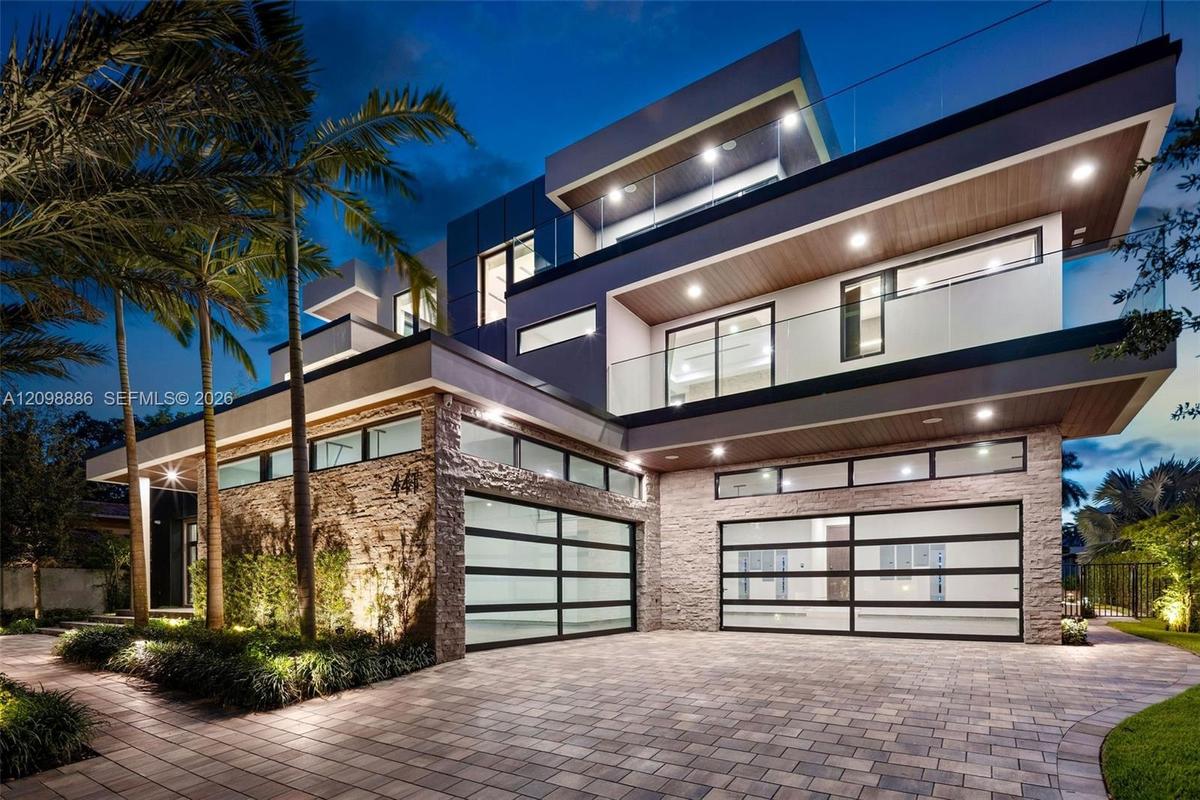 Contemporary multi-level exterior featuring stacked stone masonry, horizontal glass panel garage doors, glass-railed balconies, and wood-finish soffit