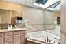 Custom wooden vanity with dual recessed-mirror cabinets, corner jetted tub, wall-to-wall tiling with decorative trim, and an expansive overhead skylig