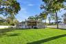 Expansive rear yard featuring a manicured lawn, perimeter white privacy fencing, mature trees, and a multi-windowed architectural exterior with a rais