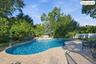 Free-form swimming pool featuring a rock waterfall feature and expansive stone paver decking, surrounded by mature trees and perimeter fencing (covere