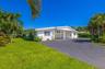 Single-story residence featuring white exterior cladding, a covered carport, and asphalt driveway, surrounded by mature palm trees and landscaped grou