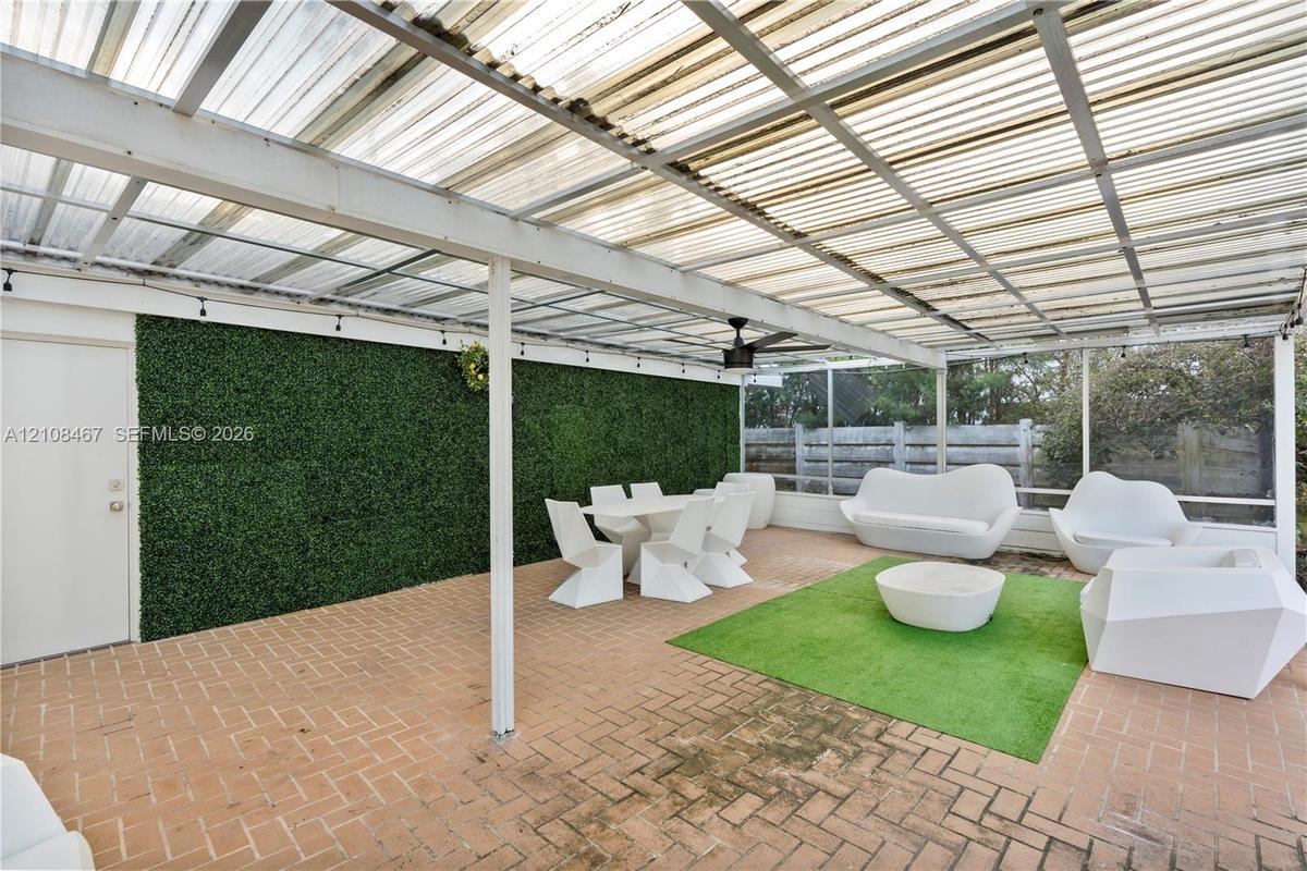 Covered patio featuring a corrugated translucent roof, brick paver flooring, green decorative privacy wall, ceiling fan, and white structural support 