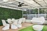 Covered patio featuring a brick-paved floor, artificial grass inlay, a matte black ceiling fan, string lighting, and a green hedge wall