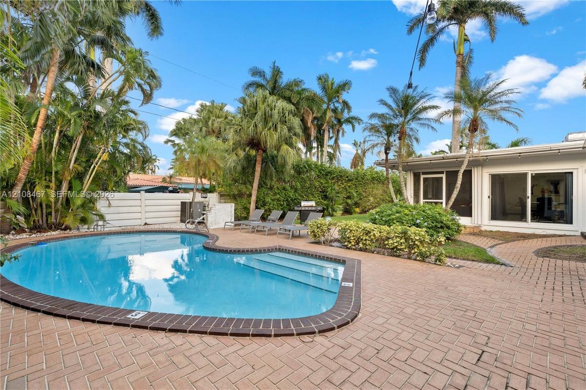 Free-form swimming pool with a dark-tiled border, expansive herringbone-pattern brick paver patio, established palm trees, and perimeter privacy fenci