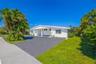 Single-story exterior featuring white stucco cladding, a paved driveway, a covered entrance, horizontal louvered windows, and mature palm trees