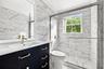 Dark navy vanity cabinet with metallic hardware, white countertop, large-format veined tile wall surfacing, glass-enclosed shower, and framed vanity m