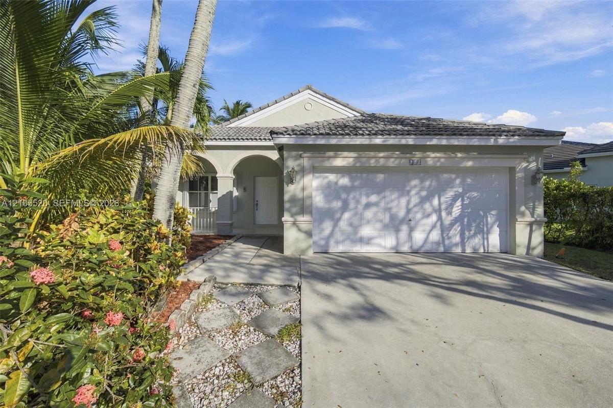 Single-story exterior featuring a concrete tile roof, arched entryway, and two-car garage, complemented by a stone paver walkway and established tropi