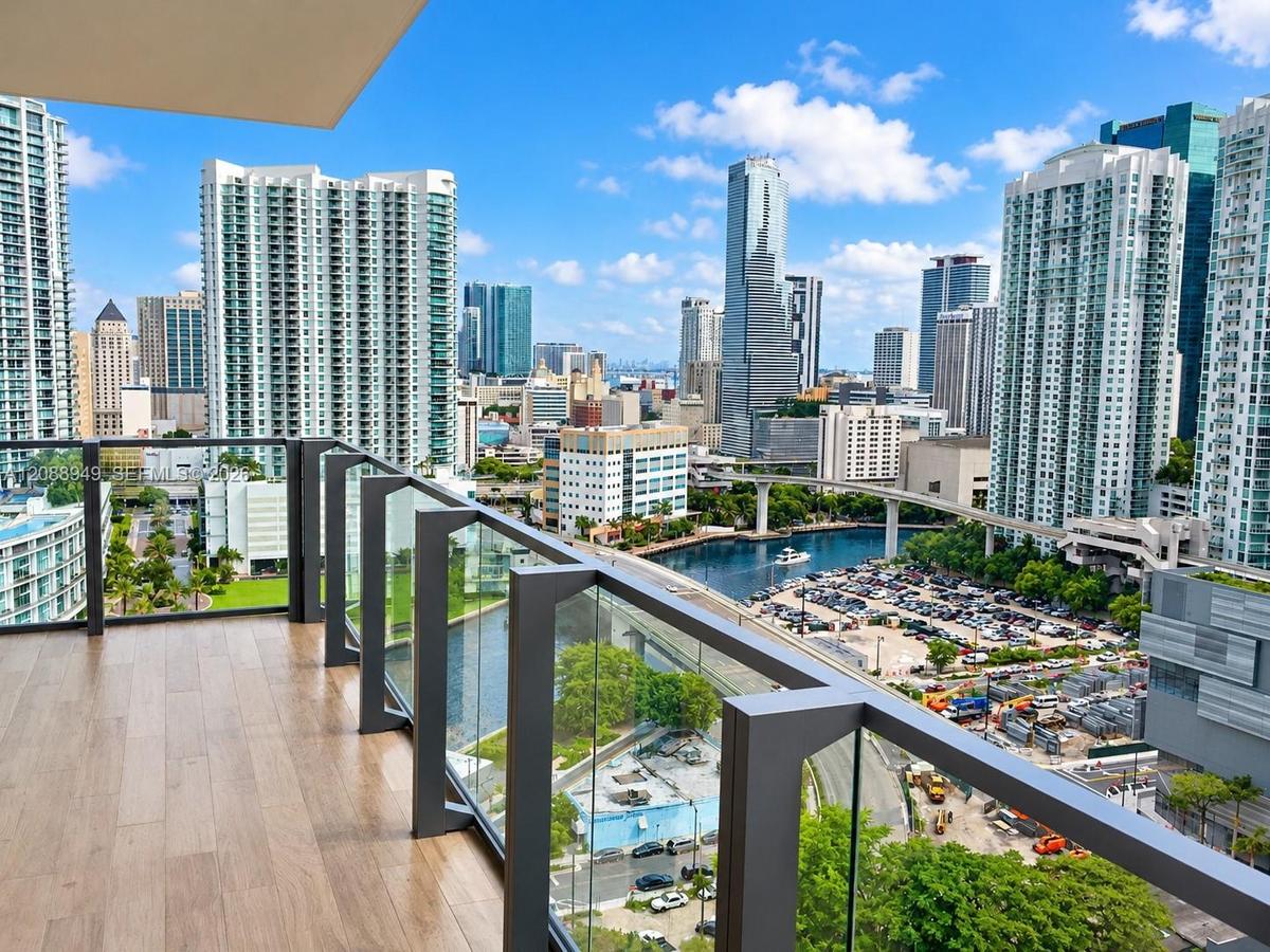 Private balcony featuring wood-finish flooring and glass panel railings with dark metal framing, overlooking an urban cityscape and waterway